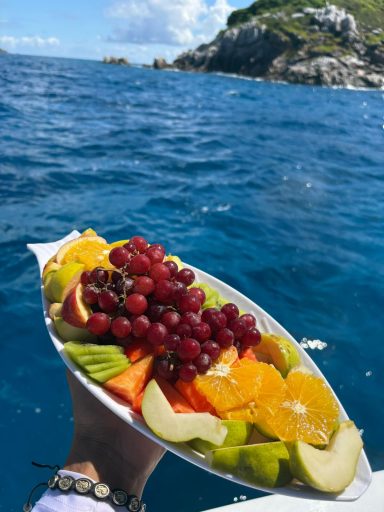 A colorful plate of fresh fruit — oranges, grapes, papaya, kiwi, and guava — held above the sea after a diving trip.