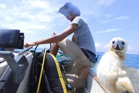 A smiling man on a boat sits next to a large white seabird resting calmly on the edge of the deck.