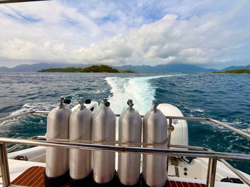 Several scuba tanks lined up on the back of a boat as it leaves the islands, creating a white wake on the sea.