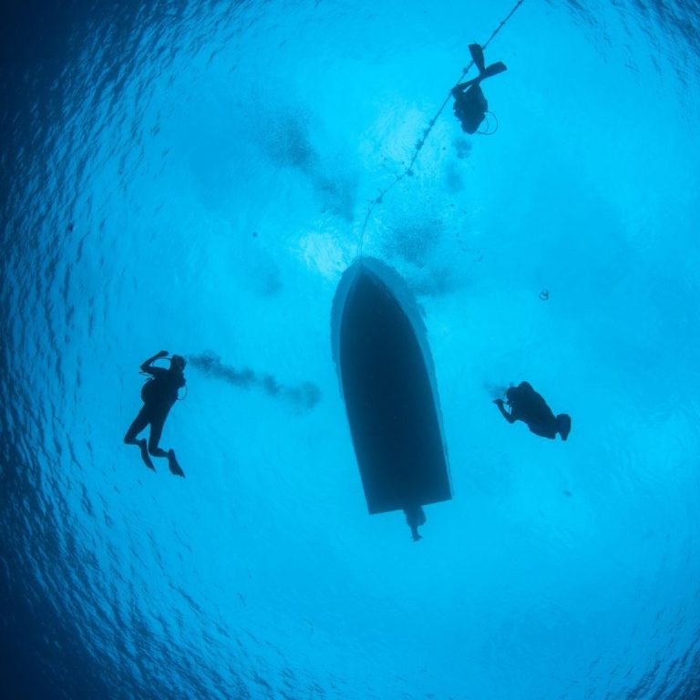 Scuba Divers Under the Boat – Safety Stop at Eden Island, Seychelles Underwater view of three scuba divers ascending beneath a boat in clear blue water.