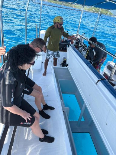Divers sit on a glass-bottom dive boat, looking through the transparent panel at the clear blue sea below.